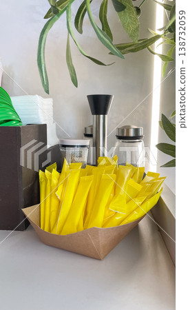 Yellow sugar sticks in a craft paper tray at a modern cafe condiment station with green plant leaves 138732059