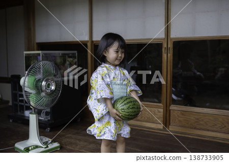 一位身穿浴衣的女孩手持西瓜，站在一棟老式日式房屋的陽台上。 138733905