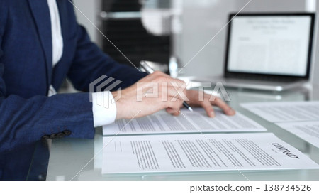 Close-up view of a business person in a professional office setting, reviewing and signing a legal contract document. Hands holding a pen over paperwork during corporate deal negotiations Close-up view of a business person in a professional office setting, reviewing and signing a legal contract document. Hands holding a pen over paperwork during corporate deal negotiations 138734526
