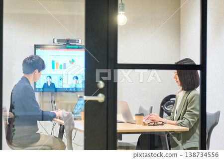 一名男子和一名女子在玻璃牆會議室與遠端團隊進行網路會議。 一名男子和一名女子在玻璃牆會議室與遠端團隊進行網路會議。 138734556