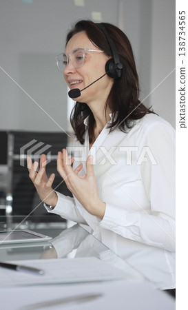 Businesswoman in a white shirt and headset leading a video call from a modern office, presenting with a tablet and gesturing as she discusses strategy and trains remote colleagues Businesswoman in a white shirt and headset leading a video call from a modern office, presenting with a tablet and gesturing as she discusses strategy and trains remote colleagues 138734565