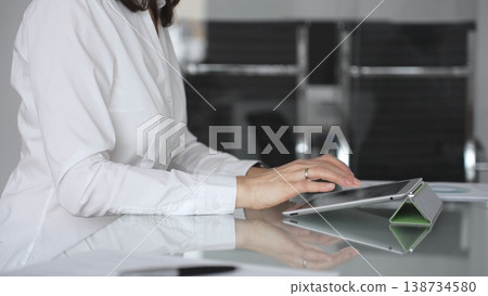 Businesswoman hands using a digital tablet while working at an office glass desk, focused on corporate operations, mobile technology, and data management in a professional workspace 138734580