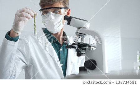Masked adult man researcher examining green botanical samples in test tube in a sterile laboratory during biological analysis. Medicine, healthcare and science concept 138734581
