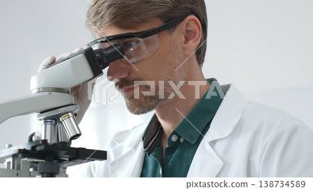 Senior man scientist wearing protective glasses and lab coat analyzing samples with a microscope in laboratory setting. Medicine, health care and science concept Senior man scientist wearing protective glasses and lab coat analyzing samples with a microscope in laboratory setting. Medicine, health care and science concept 138734589