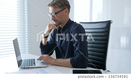 Professional businessman sitting at a sleek desk, working on a laptop and thinking about goals in modern office filled with natural light. Business people concept Professional businessman sitting at a sleek desk, working on a laptop and thinking about goals in modern office filled with natural light. Business people concept 138734591