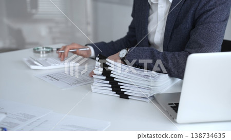 Financial professional reviewing accounts at a white desk, using a calculator and organizing a stack of documents to prepare taxes, audit reports and business financial analysis. Audit and taxes 138734635