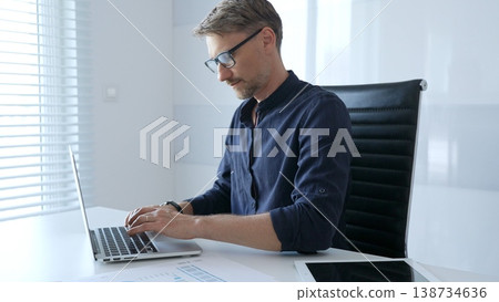 Professional businessman wearing glasses, intensely working on laptop, typing amid modern office environment with tablet and documents surrounding workspace. Business people concept 138734636