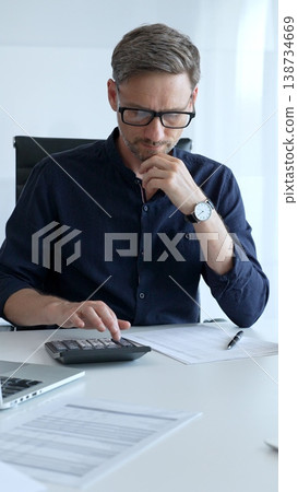 Businessman sitting at a sleek desk, working on a laptop and thinking in a bright, modern office filled with natural light. Business people concept 138734669