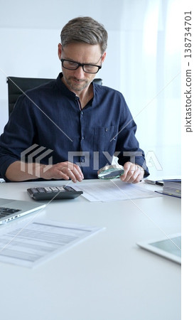 Financial analyst reviewing paperwork, using calculator and magnifying glass while examining corporate budget reports in modern workspace. Audit and taxes, business concept Financial analyst reviewing paperwork, using calculator and magnifying glass while examining corporate budget reports in modern workspace. Audit and taxes, business concept 138734701