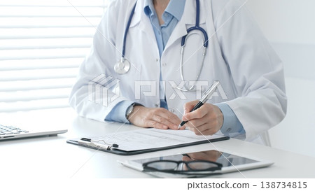 Female doctor documents patient information while working with digital devices at minimalist white workstation in medical office. Medicine and healthcare concept Female doctor documents patient information while working with digital devices at minimalist white workstation in medical office. Medicine and healthcare concept 138734815