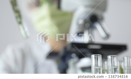 Close up of test tubes with a scientist analyzing plant samples in test tubes with a microscope in a laboratory at the background. Medicine, health care and science concept 138734816