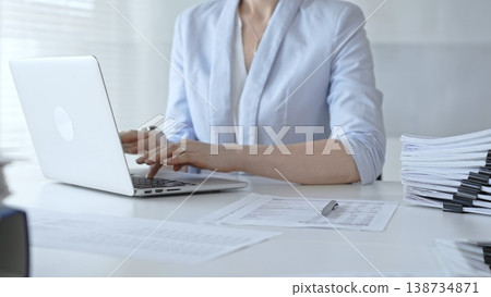 Professional woman entering and analyzing financial data on a laptop amid paperwork and binders on a tidy white desk. Audit and taxes Professional woman entering and analyzing financial data on a laptop amid paperwork and binders on a tidy white desk. Audit and taxes 138734871
