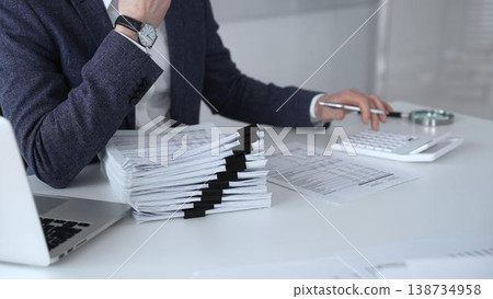 Financial professional reviewing accounts at a white desk, using a calculator and organizing a stack of documents to prepare taxes, audit reports and business financial analysis. Audit and taxes Financial professional reviewing accounts at a white desk, using a calculator and organizing a stack of documents to prepare taxes, audit reports and business financial analysis. Audit and taxes 138734958