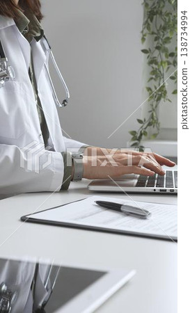 Doctor woman wearing white lab coat over green shirt, and stethoscope, is typing on laptop, documenting patient information. Medicine and health care concept Doctor woman wearing white lab coat over green shirt, and stethoscope, is typing on laptop, documenting patient information. Medicine and health care concept 138734994