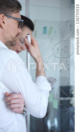 Business professionals collaborating on a transparent glass board, drawing diagrams and analyzing ideas for a new project while discussing strategy, innovation, and teamwork 138735018