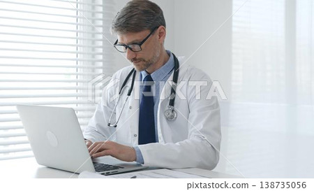 A male doctor in glasses and a lab coat with a stethoscope is typing on a laptop keyboard while sitting at a table in a bright medical office A male doctor in glasses and a lab coat with a stethoscope is typing on a laptop keyboard while sitting at a table in a bright medical office 138735056