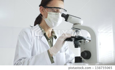 Female scientist wearing a lab coat, face mask, and safety glasses carefully adjusts a microscope, conducting research in a brightly lit laboratory setting. Medicine, healthcare and science concept Female scientist wearing a lab coat, face mask, and safety glasses carefully adjusts a microscope, conducting research in a brightly lit laboratory setting. Medicine, healthcare and science concept 138735065
