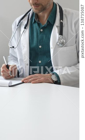 Doctor man wearing lab coat over green shirt, and stethoscope is writing medical prescription or filling medical documents sitting at office desk. Medicine and health care Doctor man wearing lab coat over green shirt, and stethoscope is writing medical prescription or filling medical documents sitting at office desk. Medicine and health care 138735087