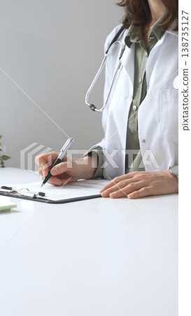 Professional female doctor wearing white lab coat and stethoscope documenting patient information on clipboard near laptop in clinic workspace. Medicine and health care Professional female doctor wearing white lab coat and stethoscope documenting patient information on clipboard near laptop in clinic workspace. Medicine and health care 138735127