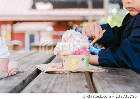 一個男孩坐在戶外長椅上,用杯子吃冰淇淋,他的手露出了這樣的表情。 一個男孩坐在戶外長椅上,用杯子吃冰淇淋,他的手露出了這樣的表情。 138735756