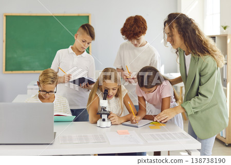 Teacher observes how girl student looks into microscope during biology lesson in primary school. 138738190