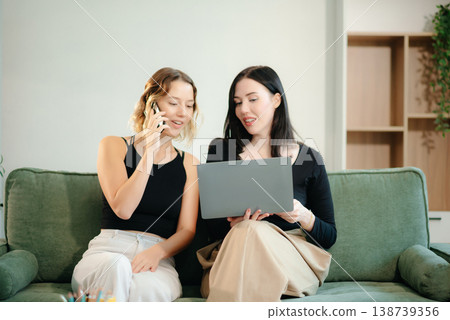 Two women collaborating on a project at home, brainstorming with a laptop and notebook in a cozy setting. Two women collaborating on a project at home, brainstorming with a laptop and notebook in a cozy setting. 138739356