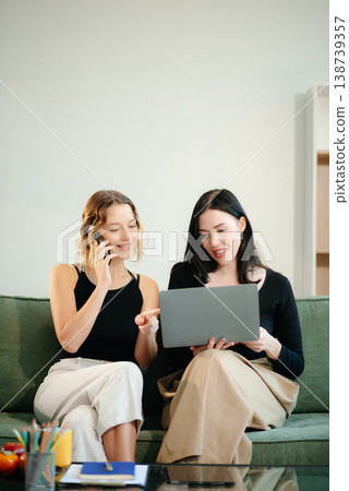 Two women collaborating on a project at home, brainstorming with a laptop and notebook in a cozy setting. Two women collaborating on a project at home, brainstorming with a laptop and notebook in a cozy setting. 138739357