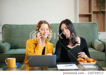 Two women collaborating on a project at home, brainstorming with a laptop and notebook in a cozy setting. Two women collaborating on a project at home, brainstorming with a laptop and notebook in a cozy setting. 138739517
