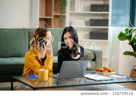 Two women collaborating on a project at home, brainstorming with a laptop and notebook in a cozy setting. Two women collaborating on a project at home, brainstorming with a laptop and notebook in a cozy setting. 138739519