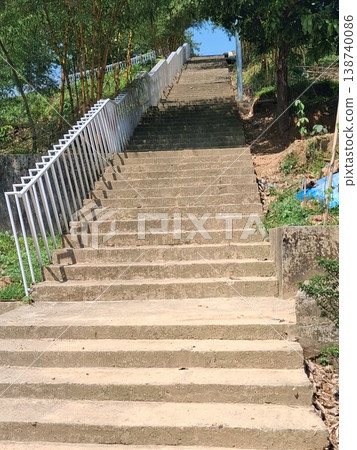 Concrete Outdoor Stairs With White Railing Rising Uphill Through Green Trees and Sunlit Pathway 138740086