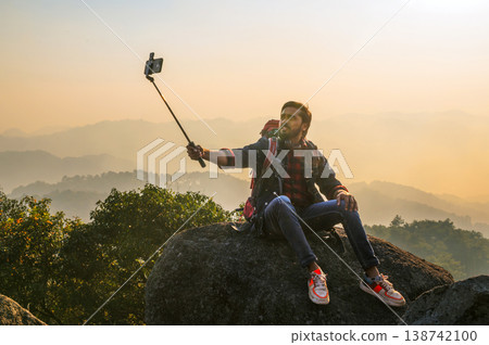 Man sits atop rocky outcrop, capturing sunrise with selfie stick Man sits atop rocky outcrop, capturing sunrise with selfie stick 138742100
