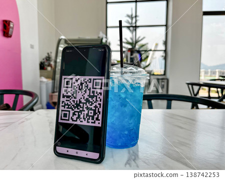 Smartphone displaying a QR code for digital payment placed on a table next to a blue drink in a cafe. Cashless transaction, mobile banking, and contactless technology in modern lifestyle setting 138742253