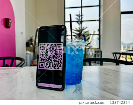 Smartphone displaying a QR code for digital payment placed on a table next to a blue drink in a cafe. Cashless transaction, mobile banking, and contactless technology in modern lifestyle setting Smartphone displaying a QR code for digital payment placed on a table next to a blue drink in a cafe. Cashless transaction, mobile banking, and contactless technology in modern lifestyle setting 138742254
