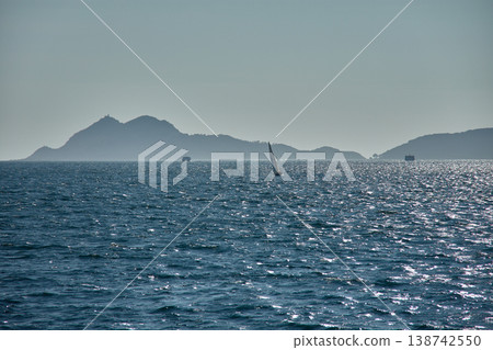 A sailboat sails at sunset through the Vigo Estuary, with the silhouettes of the Cies Islands outlined on the horizon and the shimmering waters. A sailboat sails at sunset through the Vigo Estuary, with the silhouettes of the Cies Islands outlined on the horizon and the shimmering waters. 138742550