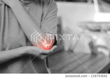 Monochrome photo of woman suffering from severe elbow pain, holding her arm with a red glowing anatomical bone graphic overlay showing joint inflammation, arthritis, or injury in medical concept. Monochrome photo of woman suffering from severe elbow pain, holding her arm with a red glowing anatomical bone graphic overlay showing joint inflammation, arthritis, or injury in medical concept. 138743682