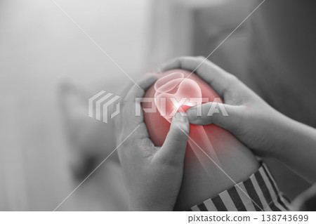 Monochrome photo of hands holding a sore knee with red glowing anatomical bone graphic overlay showing joint inflammation, pain, or arthritis in healthcare concept and medical diagnosis. 138743699