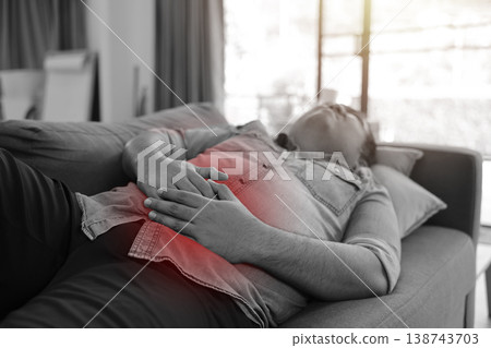 Monochrome photo of sick man lying on sofa holding his stomach with a red glowing area indicating severe abdominal pain, food poisoning, indigestion or gastric ulcer in healthcare medical concept. 138743703