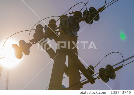 Backlit pole silhouette with electric lines, low sun creating bright flare and contrasty outlines of insulators, warm highlights and atmospheric mood photography 138743749