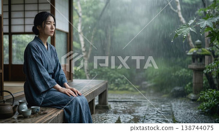 A Japanese woman in a kimono quietly relaxes on a veranda surrounded by lush greenery in the rain. 138744075