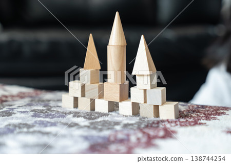 Natural wood blocks form small castle with three pointed towers as basic geometric pieces stand in ordered rows on soft fabric creating calm indoor play scene for spatial learning. 138744254