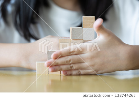 oth hands hold a wooden block tower as small fingers slowly adjust and place pieces one by one on a rug keeping the structure upright neat and stable through careful practice. oth hands hold a wooden block tower as small fingers slowly adjust and place pieces one by one on a rug keeping the structure upright neat and stable through careful practice. 138744265