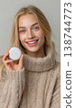 Happy young blonde woman smiling holding round moist nicotine pouch tobacco product container wearing beige knitted warm sweater 138744773