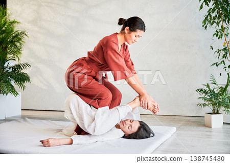 Masseuse making thai yoga massage. Woman therapist doing traditional massage treatment, stretching female patient's body. Client lying on mat, enjoying hand massage in calm atmosphere. Masseuse making thai yoga massage. Woman therapist doing traditional massage treatment, stretching female patient's body. Client lying on mat, enjoying hand massage in calm atmosphere. 138745480