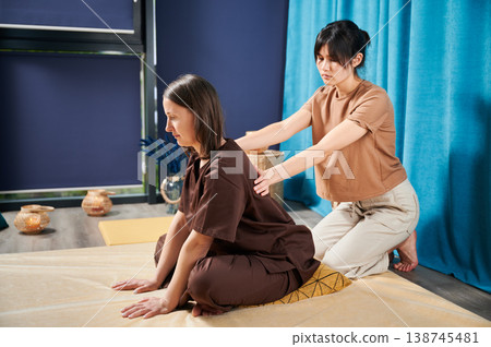 Masseuse making thai yoga massage. Woman therapist doing traditional massage treatment for female patient indoors. Client sitting on mat, enjoying hand massage in calm atmosphere. 138745481