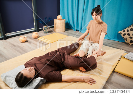 Masseuse making thai yoga massage. Woman therapist doing traditional massage treatment for female patient indoors. Client lying on mat, meditating, enjoying hand massage in calm atmosphere. 138745484