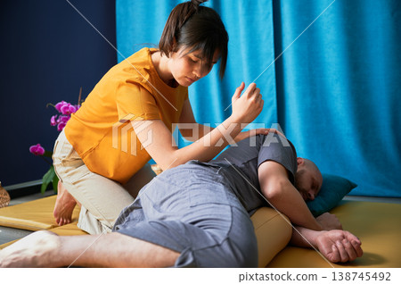 Masseuse making thai yoga massage. Woman therapist doing traditional massage treatment using elbow for male patient indoors. Client lying on mat, meditating, enjoying hand massage in calm atmosphere. 138745492