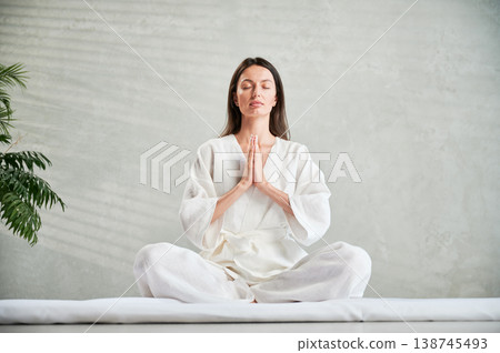 Beautiful woman in white dress sitting on mat in lotus position before Thai massage, practicing yoga, showing namaste sign. 138745493