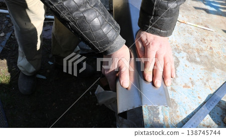 Strong male hands bending small sheet metal on an outdoor workbench, manual metalworking in a workshop, bending a piece of metal along the edge of a tabletop, human hand strength. Strong male hands bending small sheet metal on an outdoor workbench, manual metalworking in a workshop, bending a piece of metal along the edge of a tabletop, human hand strength. 138745744