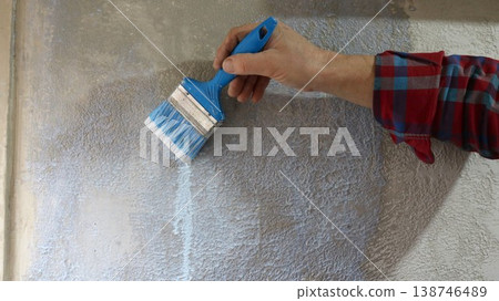 Close up of man in plaid shirt holding blue paintbrush applying liquid primer to textured grey plaster wall and priming wall of house with uneven textured plaster using paintbrush. 138746489