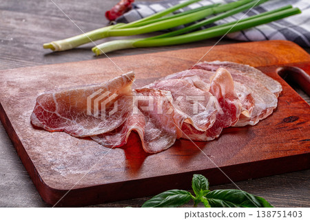 Thinly sliced cured meat, possibly prosciutto, on a wooden cutting board with green onions and basil. 138751403